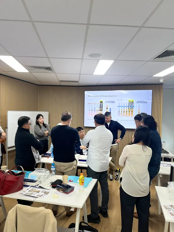 Dr JohnPaul seated at a demonstration table, explaining the mandibular drilling sequence on screen to dentists gathered around him