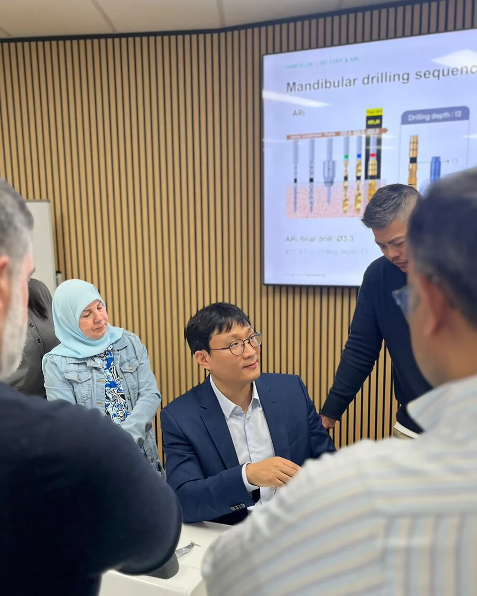 Dr JohnPaul seated at the demonstration table during his hands-on implant seminar, with participants watching and the drilling sequence on screen behind him